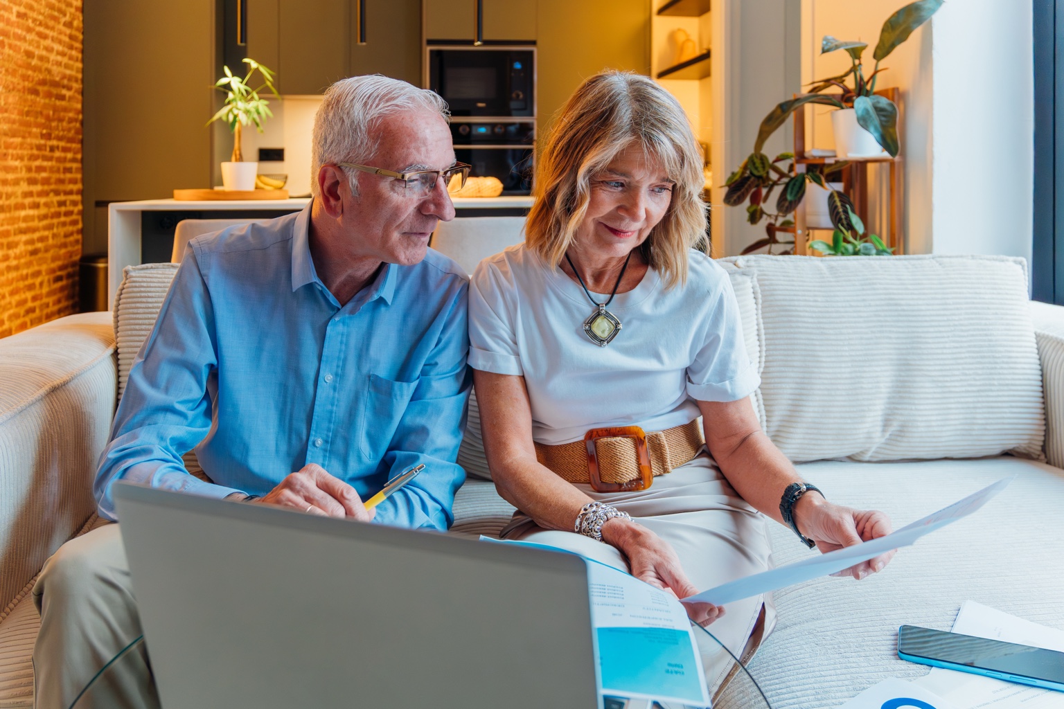 Older couple seated on a sofa reviewing printed documents beside a laptop